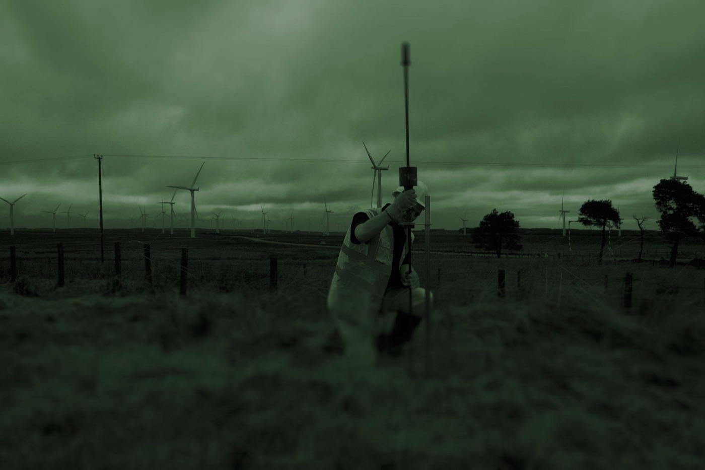 A technician in high-visibility clothing holding sampling equipment in an open field, wind turbines on the horizon.